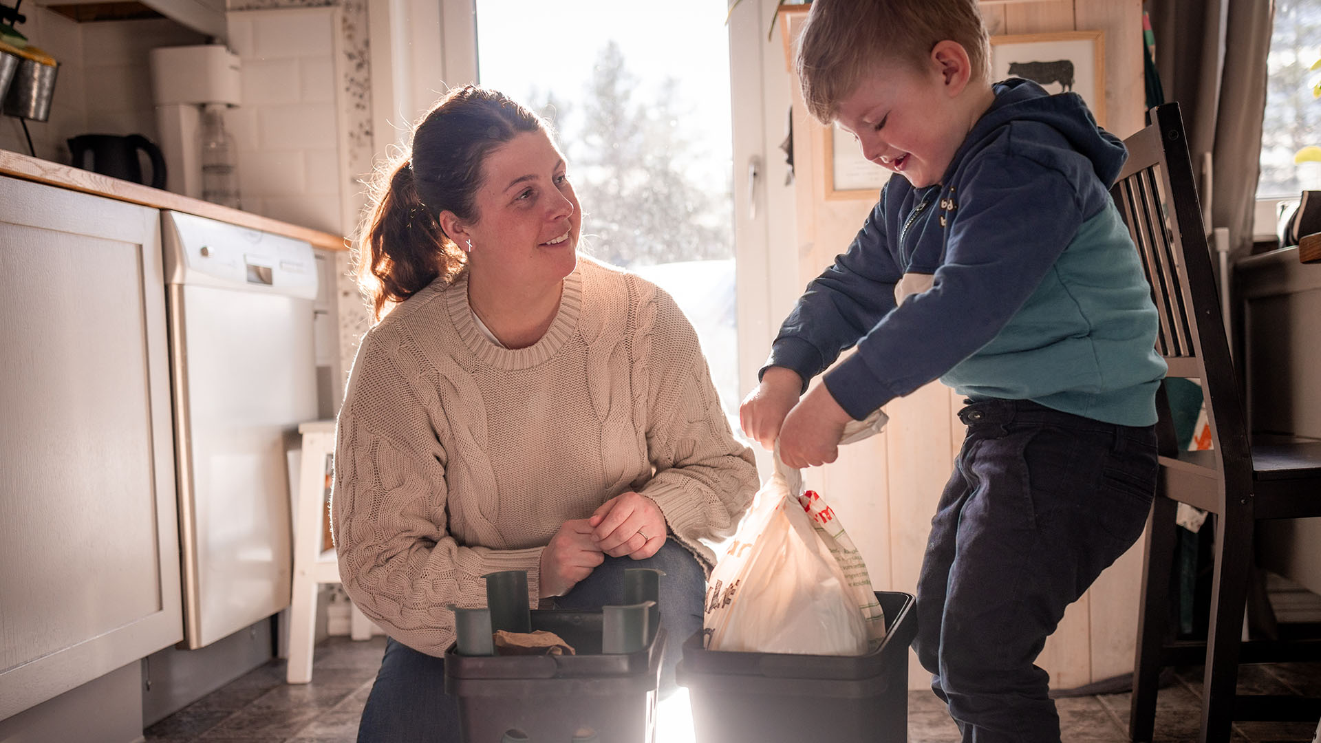 kvinna som sitter på golvet i ett kök och tittat på ett barn som knyter en soppåse.